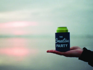 a hand holds a koozie that reads "the creative party" in front of a sunset