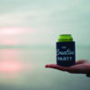 a hand holds a koozie that reads "the creative party" in front of a sunset