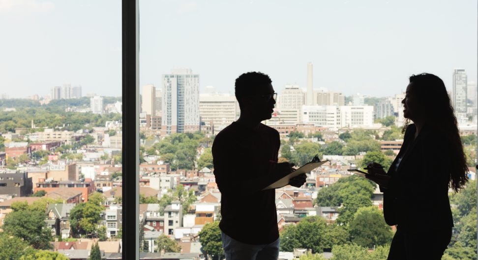 coworkers stand and talk in front of a large window overlooking a city
