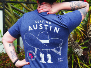 a white person with tattoos stands in front of cacti in a blue t-shirt that reads "serving Austin since 2013" and features an illustration of a figure wearing a large hat and cowboy boots