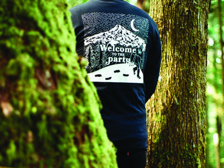 in a forest, a person's back bears a shirt with an illustration of the moon, a mountain, and sasquatch carrying a six pack of beer. it features text reading "welcome to the party"