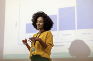 Black woman stands calmly and confidently in front of presentation deck speaking with ease