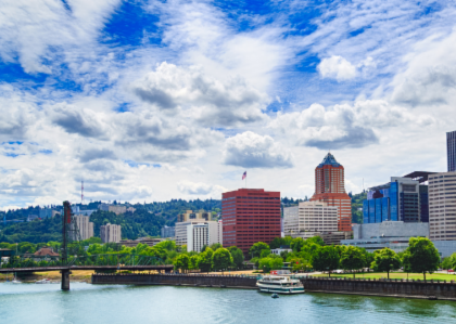 A partly-cloudy sunny day in Portland, Oregon with a view from the east side of the city of the downtown buildings collected along side the waterfront of the Columbia river.