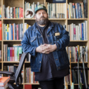 Aaron Draplin stands center in the photo leaning against a black office chair in front of a full shelf of books.