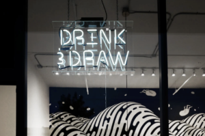Drink & Draw white neon sign hanging in a window of a brightly lit studio space.