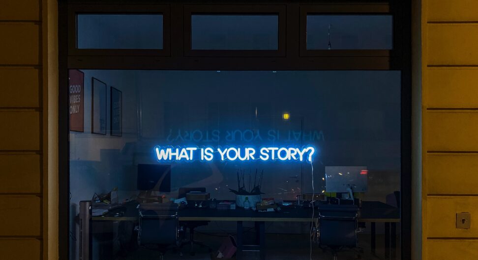 A blue neon sign hangs in the window of a store-front that reads "What is your story?"