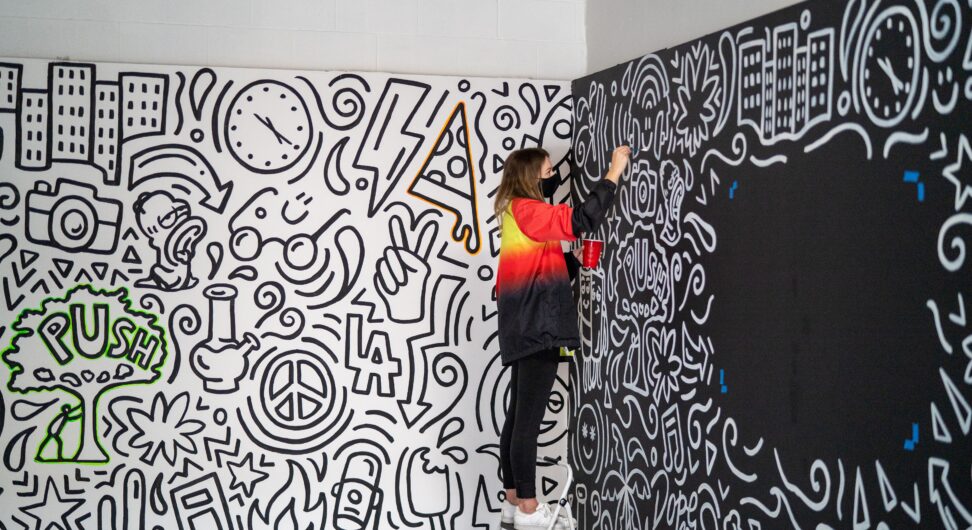 Woman stands on ladder to pain a graphic mural on a black wall.